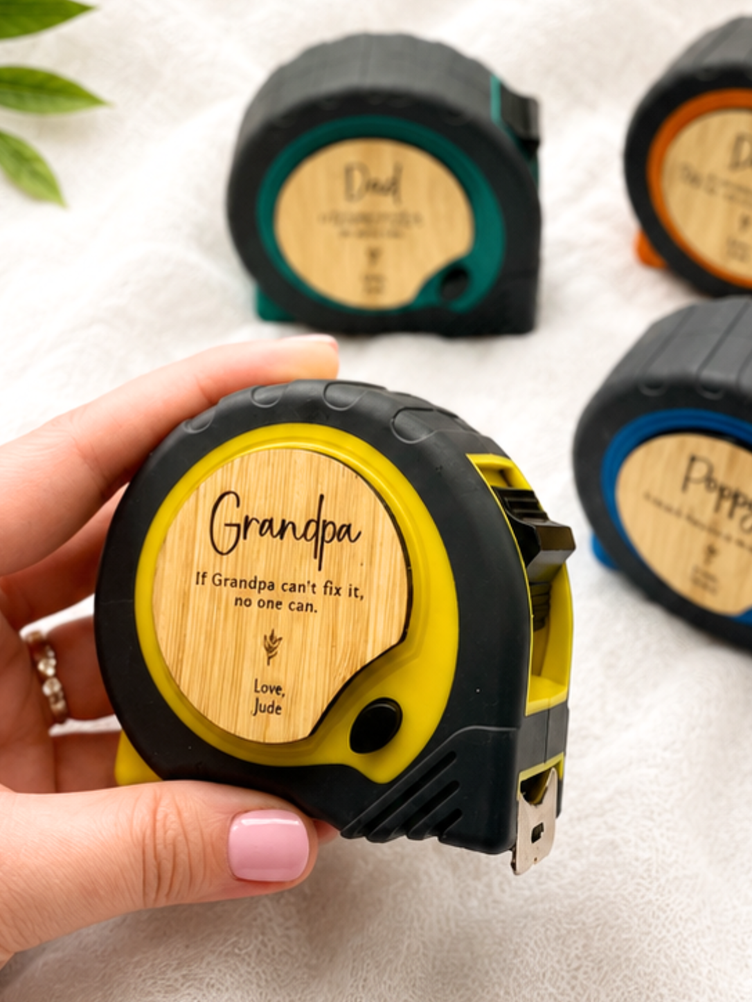 A hand holds a black and yellow retractable tape measure with a personalized bamboo-style face that reads 'Grandpa If Grandpa can't fix it, no one can. Love, Jude.