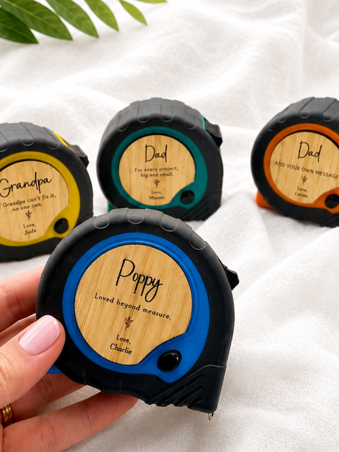 A hand holds a black tape measure with a blue rim and a bamboo-style circular face engraved with the name Poppy. Three more tape measures are blurred in the background on a white fabric surface, each with different colored rims and engraved names: Grandpa, Dad, and Dad.