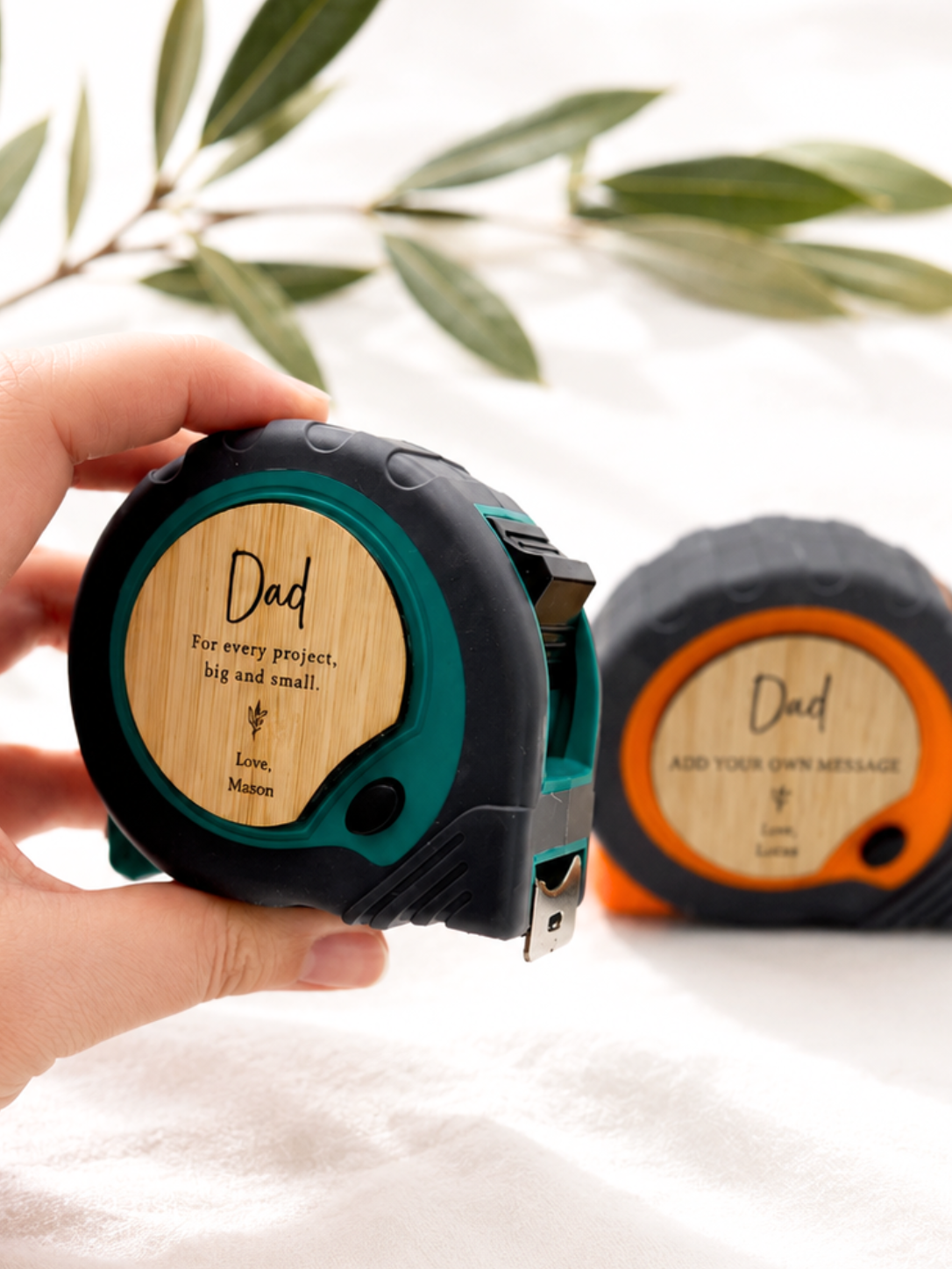 A hand holds a black and teal tape measure with a personalized bamboo insert that says 'Dad For every project, help and small.' Another tape measure sits in the background on a white fabric surface with blurred green leaves.