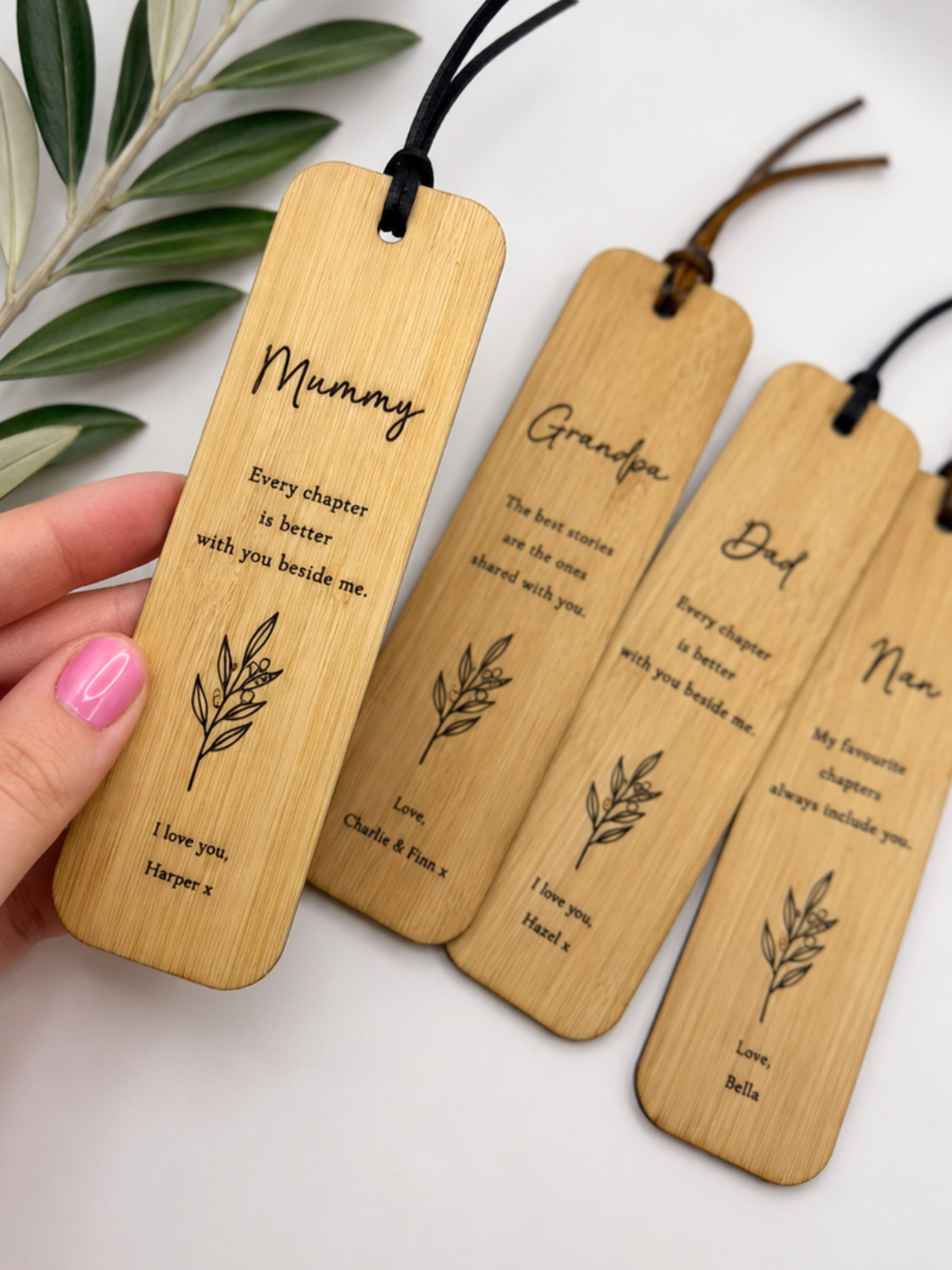 Four personalised bamboo book tags with black cords, held by a hand with pink nail polish. The tags are engraved with names (Mummy, Grandpa, Dad, and Nan) set against a white background with a green leafy branch in the corner.
