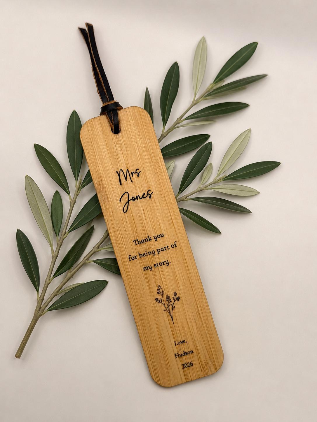 A personalized bamboo book tag with a dark brown leather loop, engraved with 'Mrs. Jones' and a thank you message, styled on an olive branch against a light beige background.