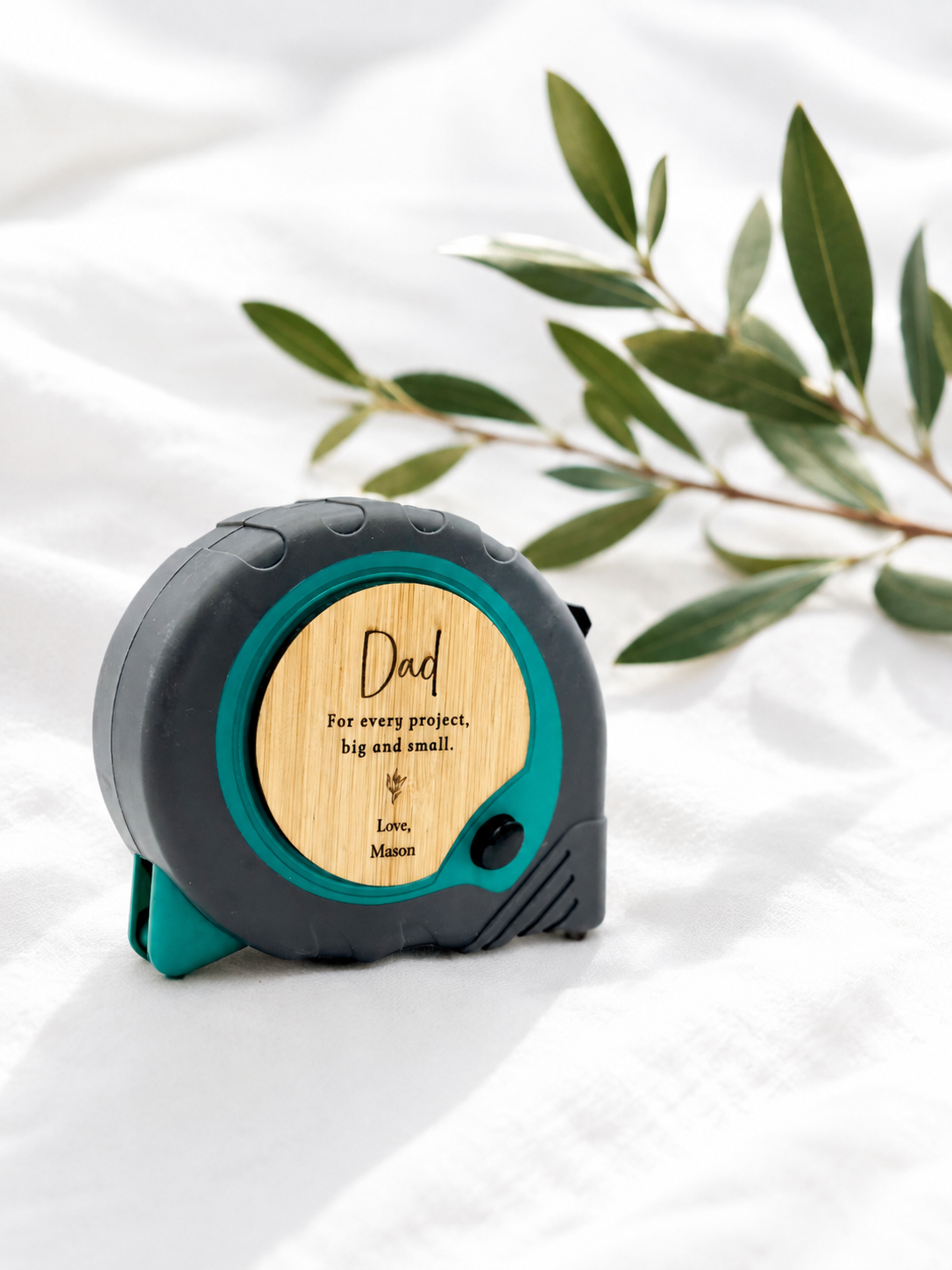 A personalized retractable tape measure with a dark gray and teal casing, featuring a bamboo wood insert engraved with 'Dad For every project, big and small.' and 'Love, Mason,' displayed on a white fabric background with green leaves.