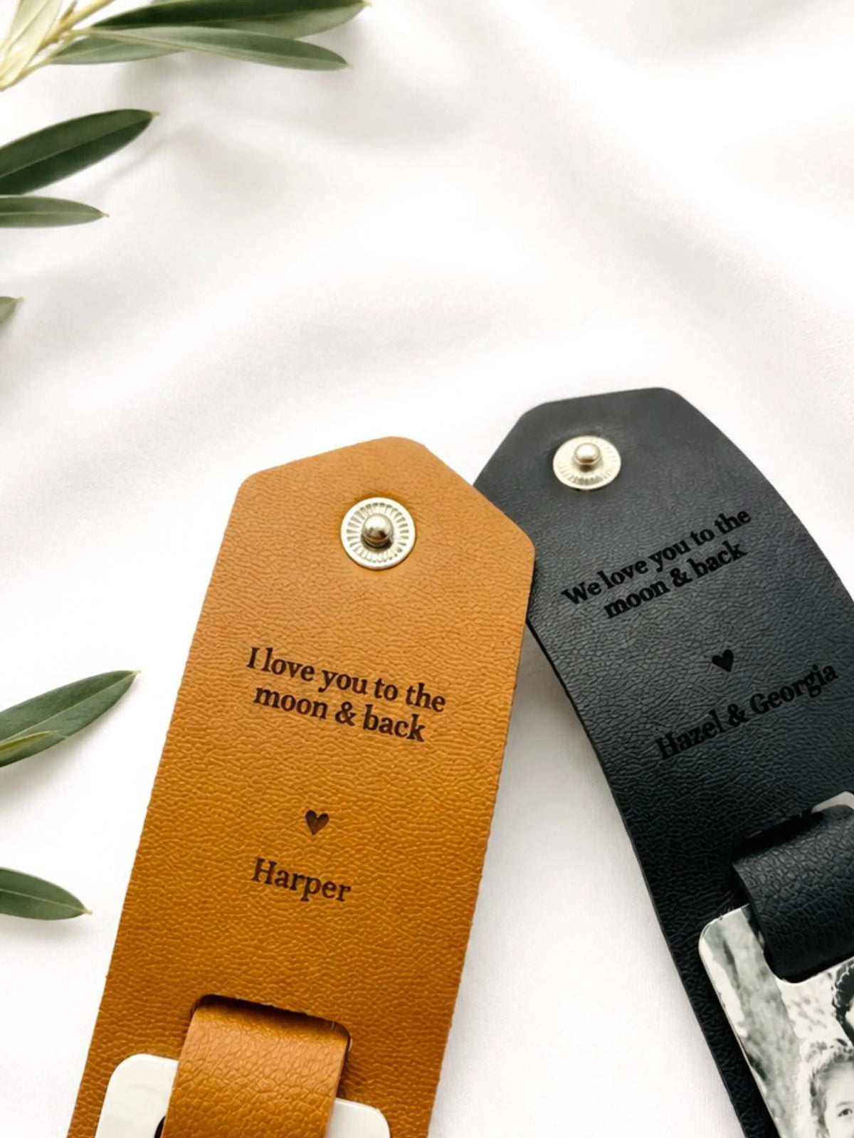 Two personalized leather keychains or tags laid on a white fabric background with olive leaves at the corner. One is tan/caramel brown with a silver-tone stud and engraved text reading 'I love you to the moon & back' and the name 'Harper.