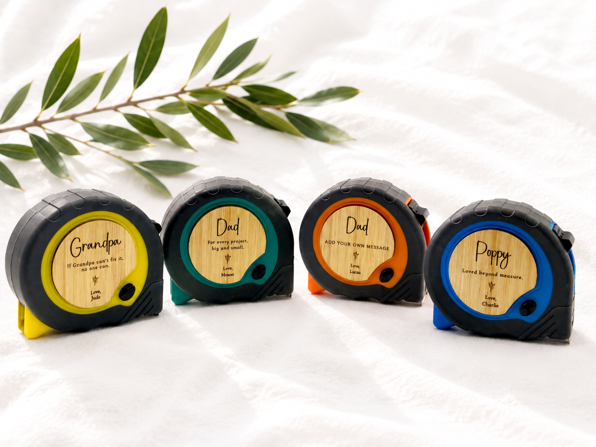Four laser level tape measures in different colours (yellow, green, orange, and blue) lined up on a white fabric background with a green leafy branch.